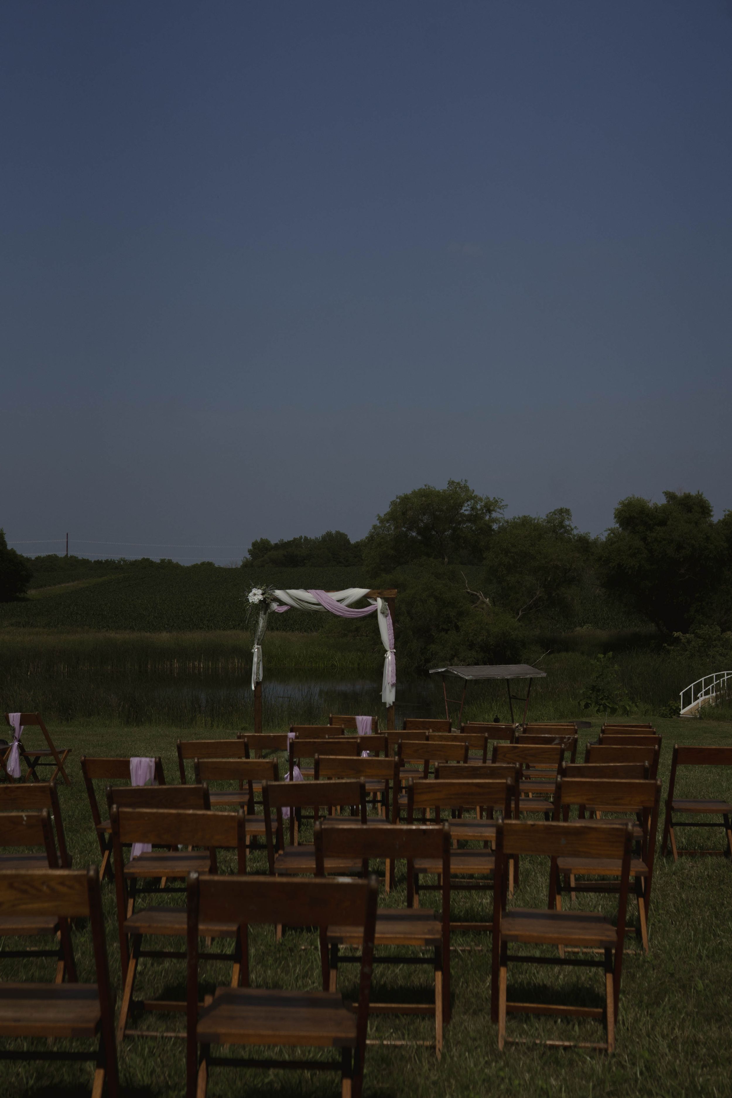 Wooden chairs set up for an outdoor ceremony with white arch and rural backdrop.