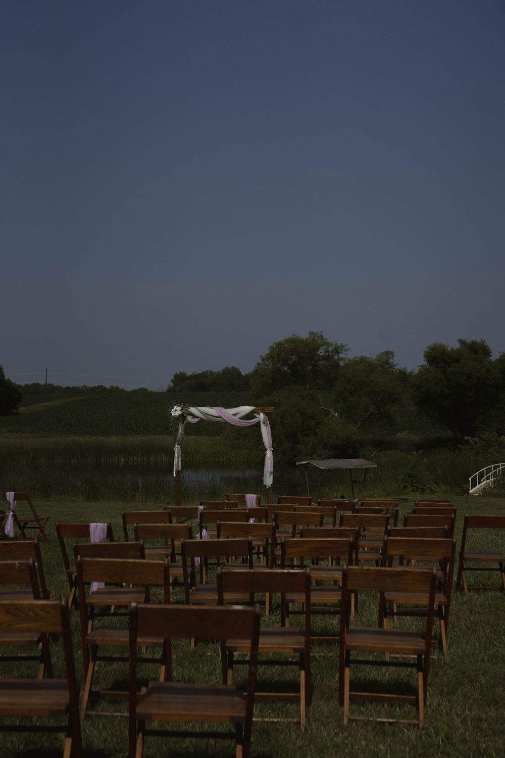 Wooden chairs set up for an outdoor ceremony with white arch and rural backdrop.