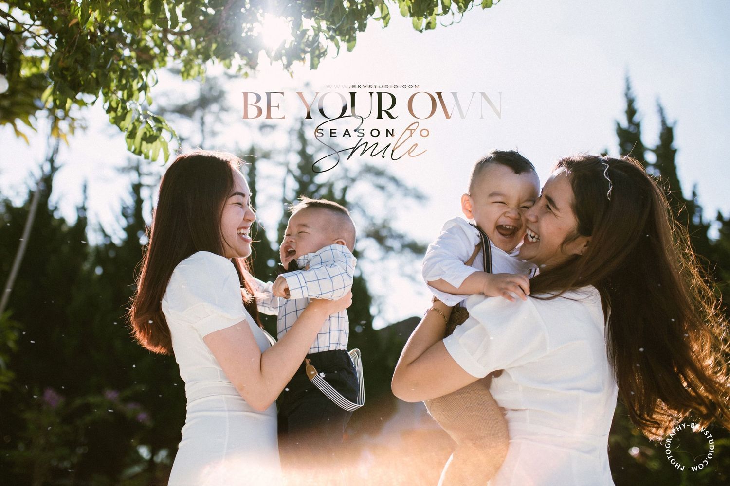 Two mothers in white dresses hold and laugh with their babies in a sunlit outdoor setting with trees.