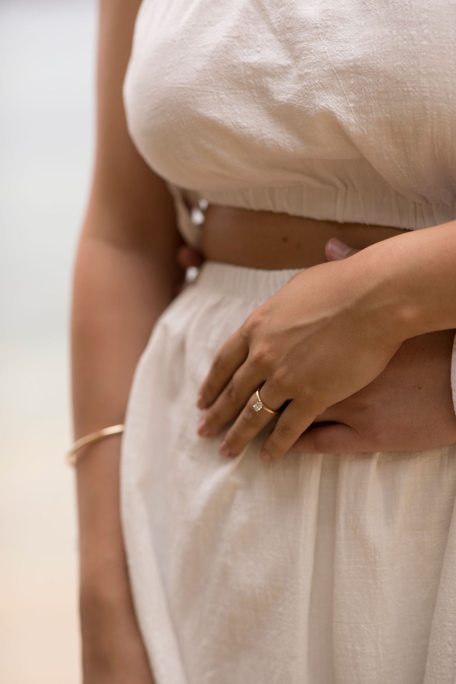 Close-up view of a hand wearing an engagement ring with a white flowing dress in soft natural lighting.