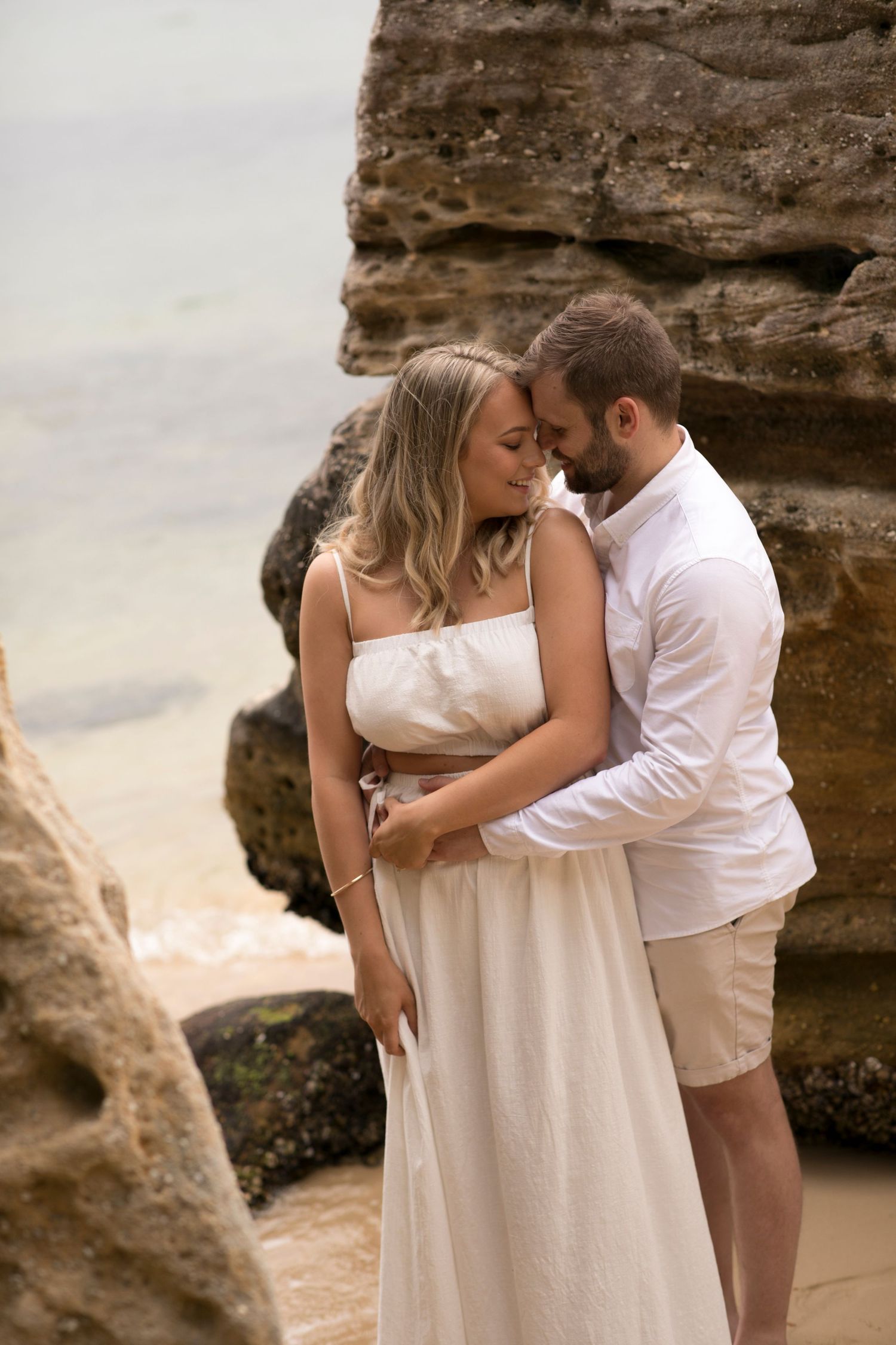 Romantic couple embraces against rocky coastal backdrop wearing matching white outfits at sunset.