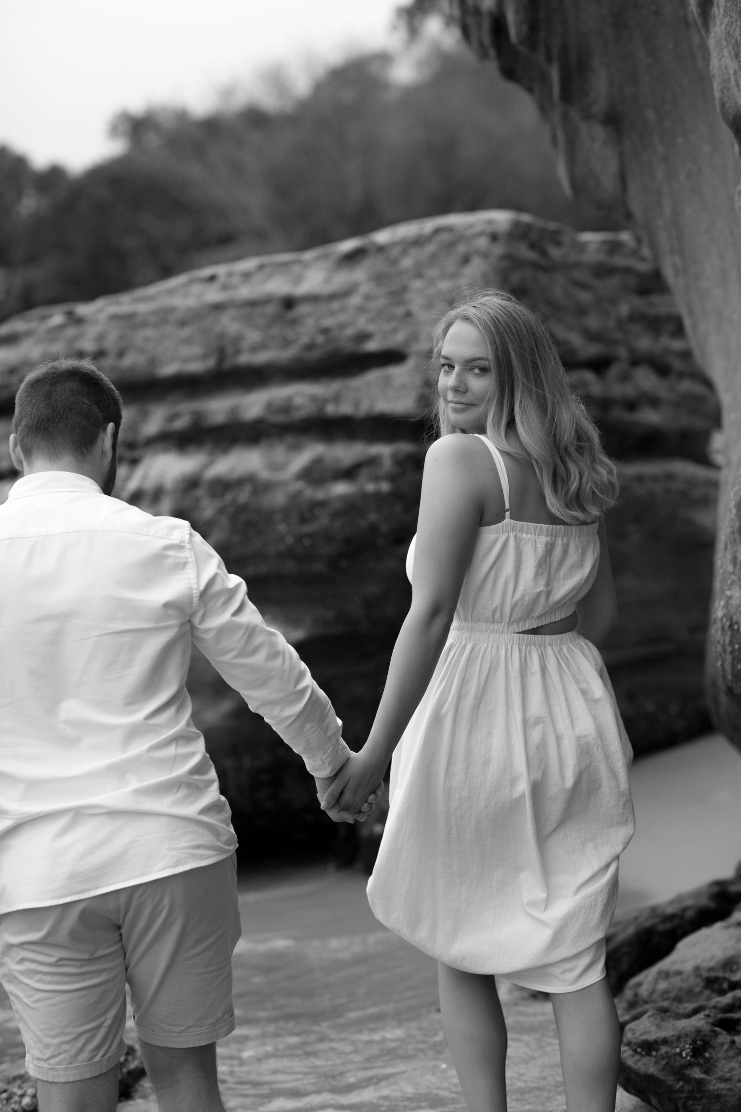 A couple wearing white outfits holds hands while walking along rocky coastal terrain in black and white.