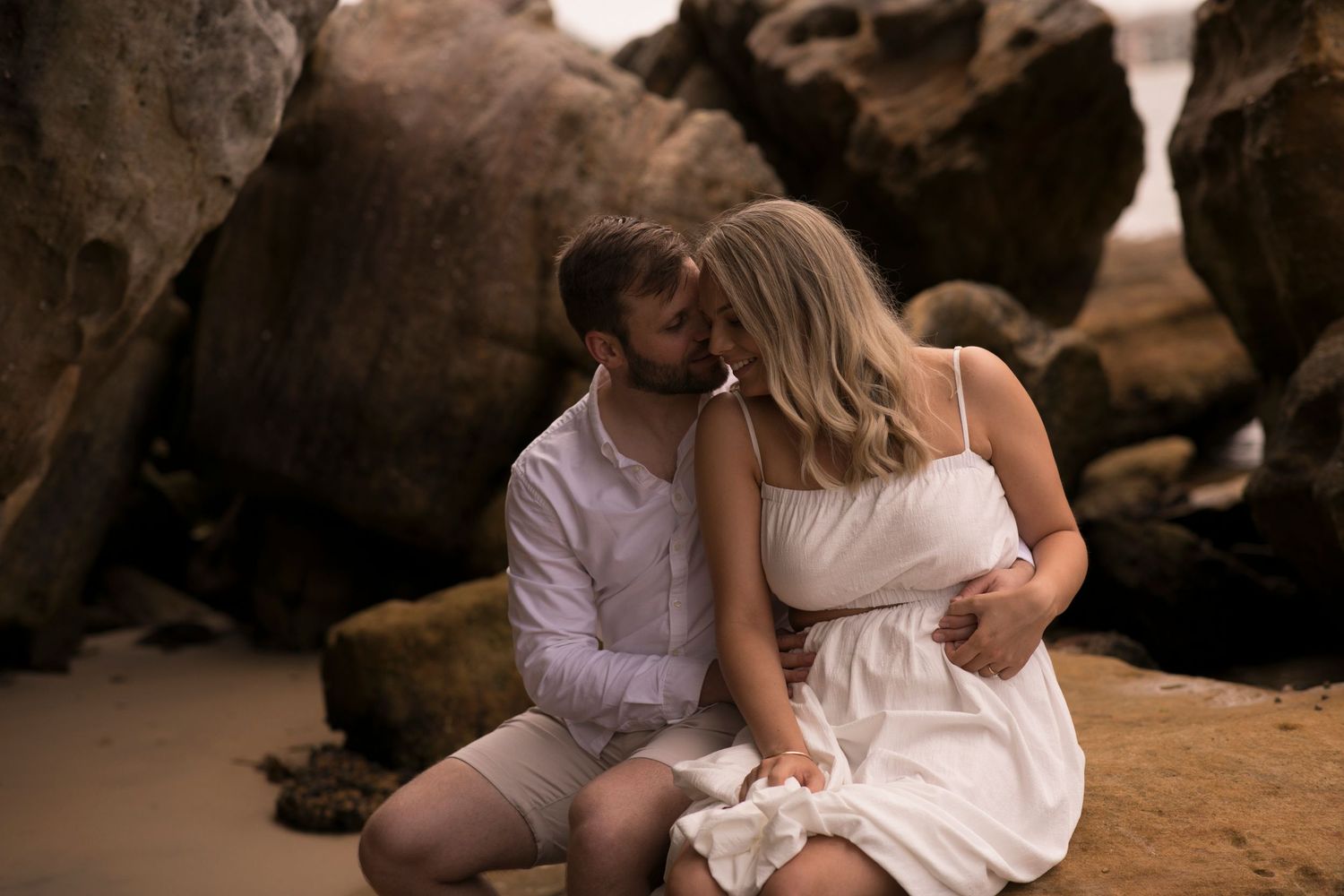 Romantic couple in white outfits sharing an intimate moment on a beach surrounded by rocky cliffs at sunset.