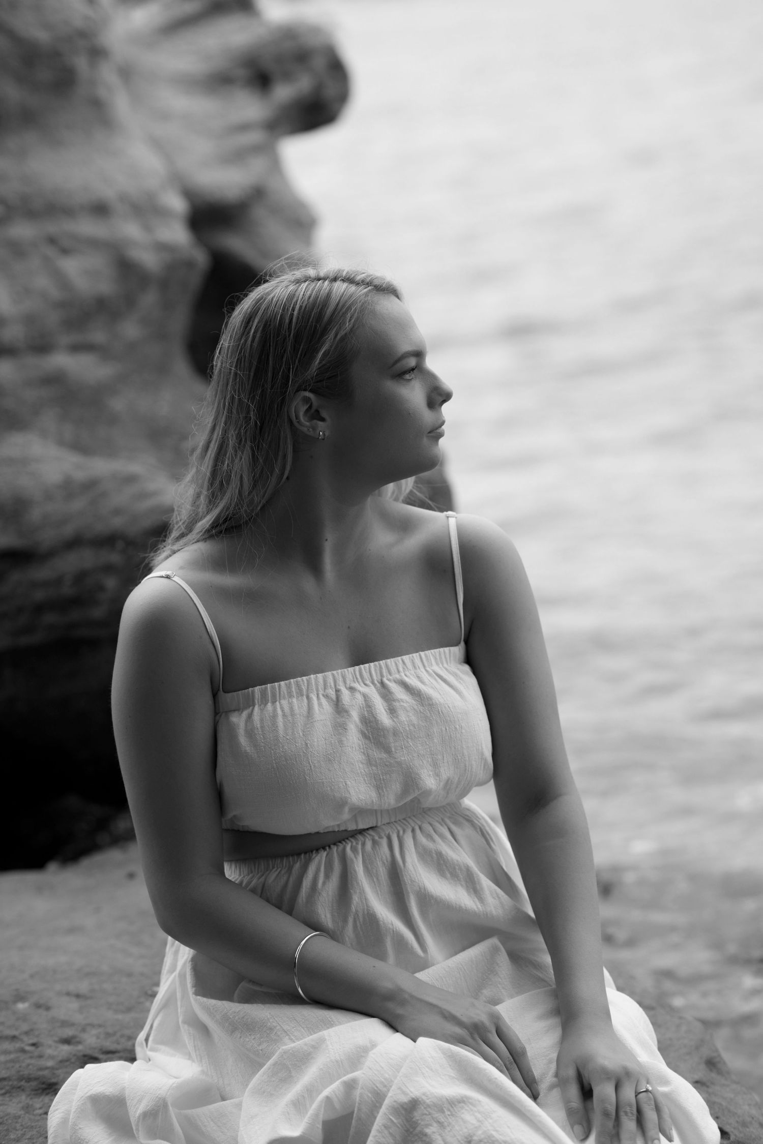 A woman in a white sundress sits thoughtfully by rocky waterfront in black and white portrait.