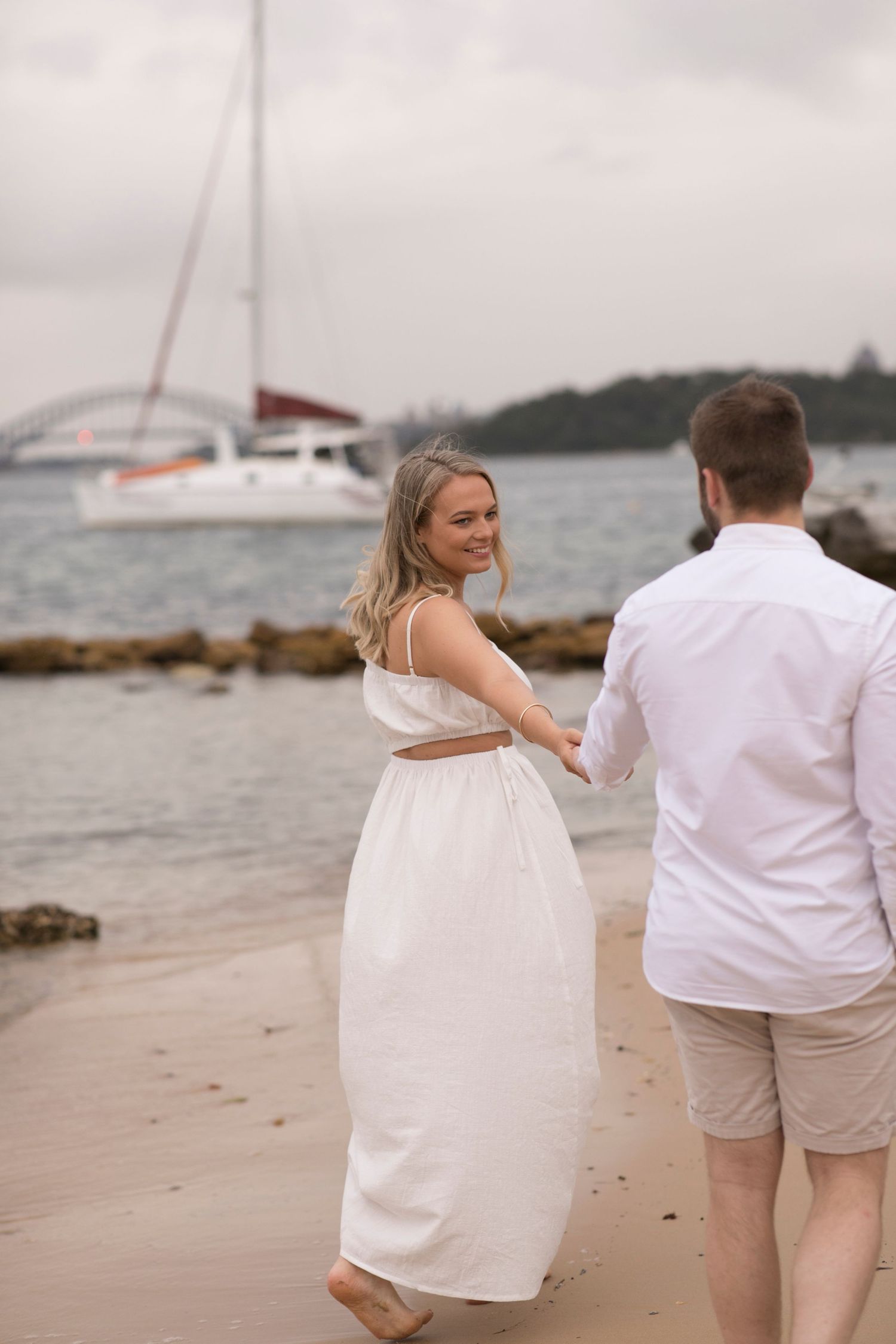 Couple in white outfits sharing a romantic moment on a beach with sailboats in the background on an overcast day.