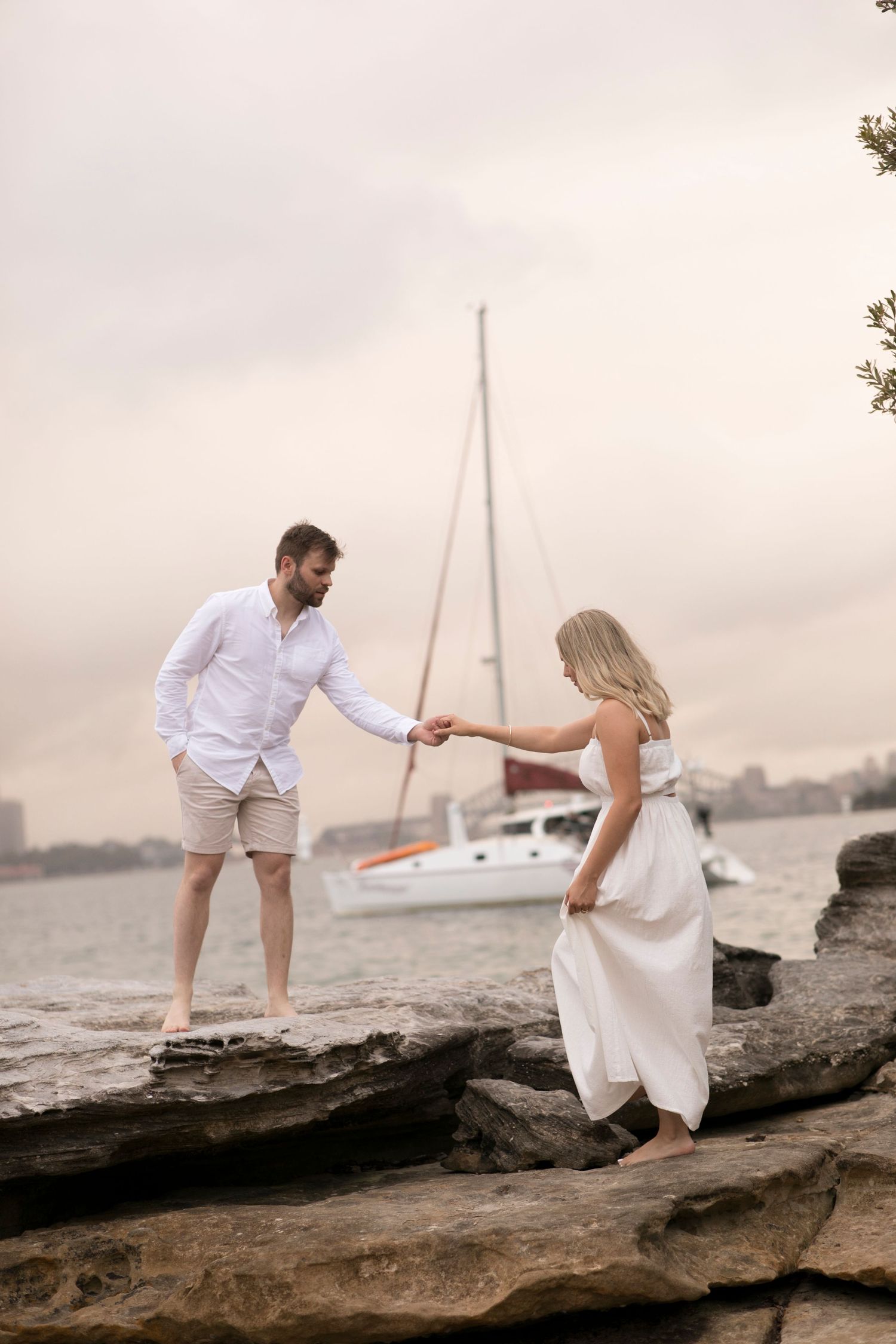 Couple holding hands on coastal rocks with sailboat in background.