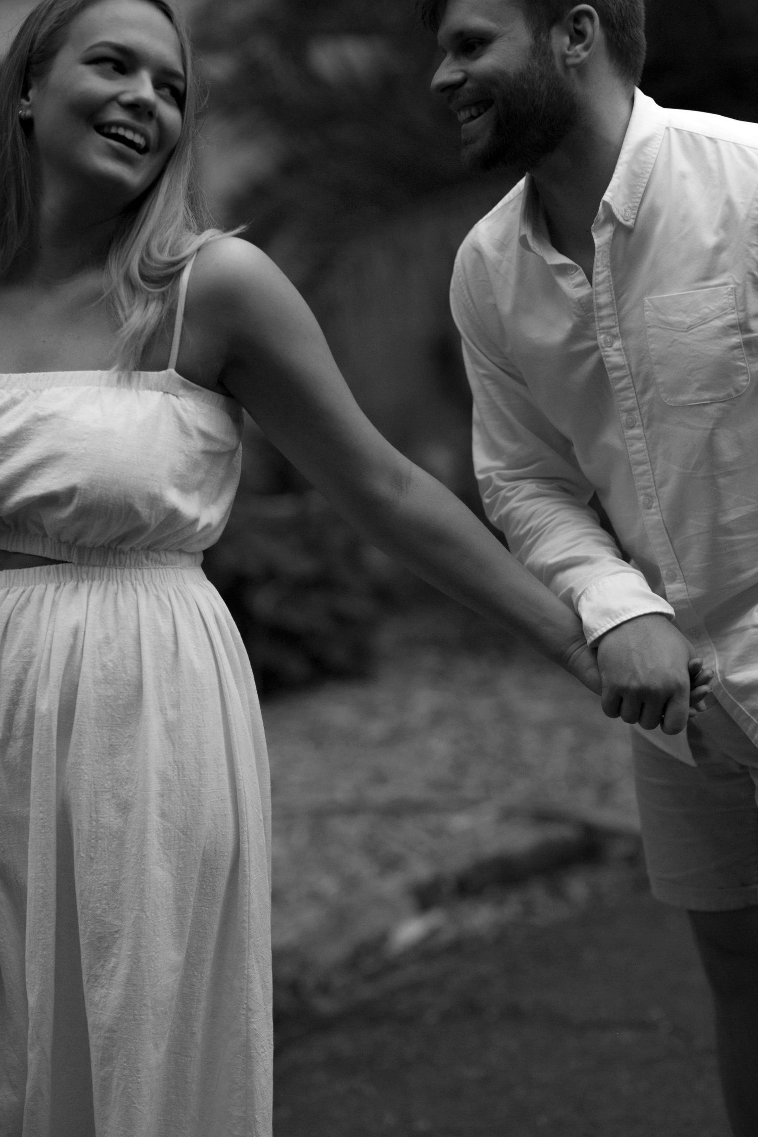 Black and white romantic photo of couple in casual white outfits holding hands and laughing in a natural setting.
