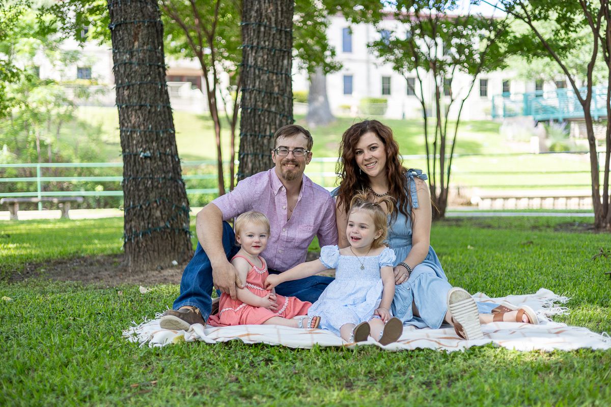 Family enjoys a picnic on a blanket under shaded trees in a grassy park on a sunny day.