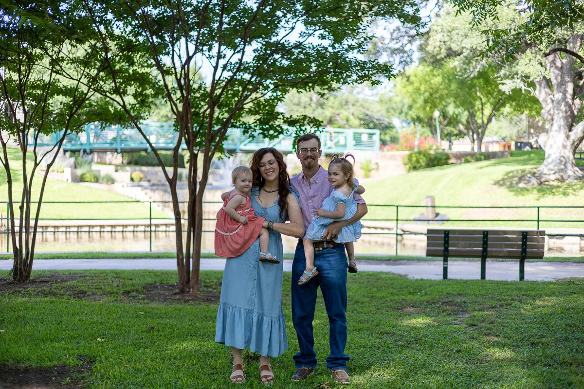 A family stands together outdoors in a park setting with green trees and benches visible in the background.