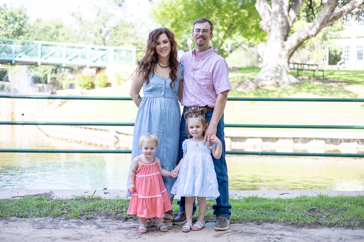Family wearing coordinated pastel outfits poses together by a waterfront railing in a park setting.