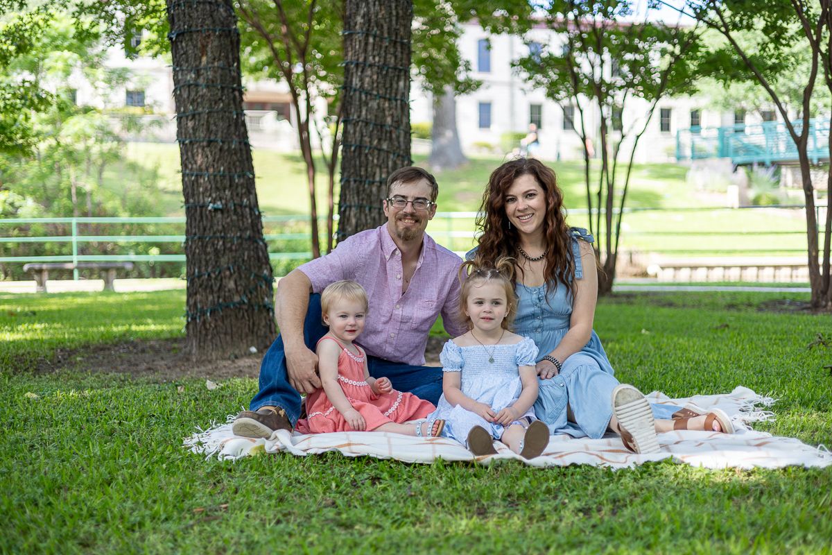 Family enjoying a summer picnic together on a blanket under shady trees in a park.