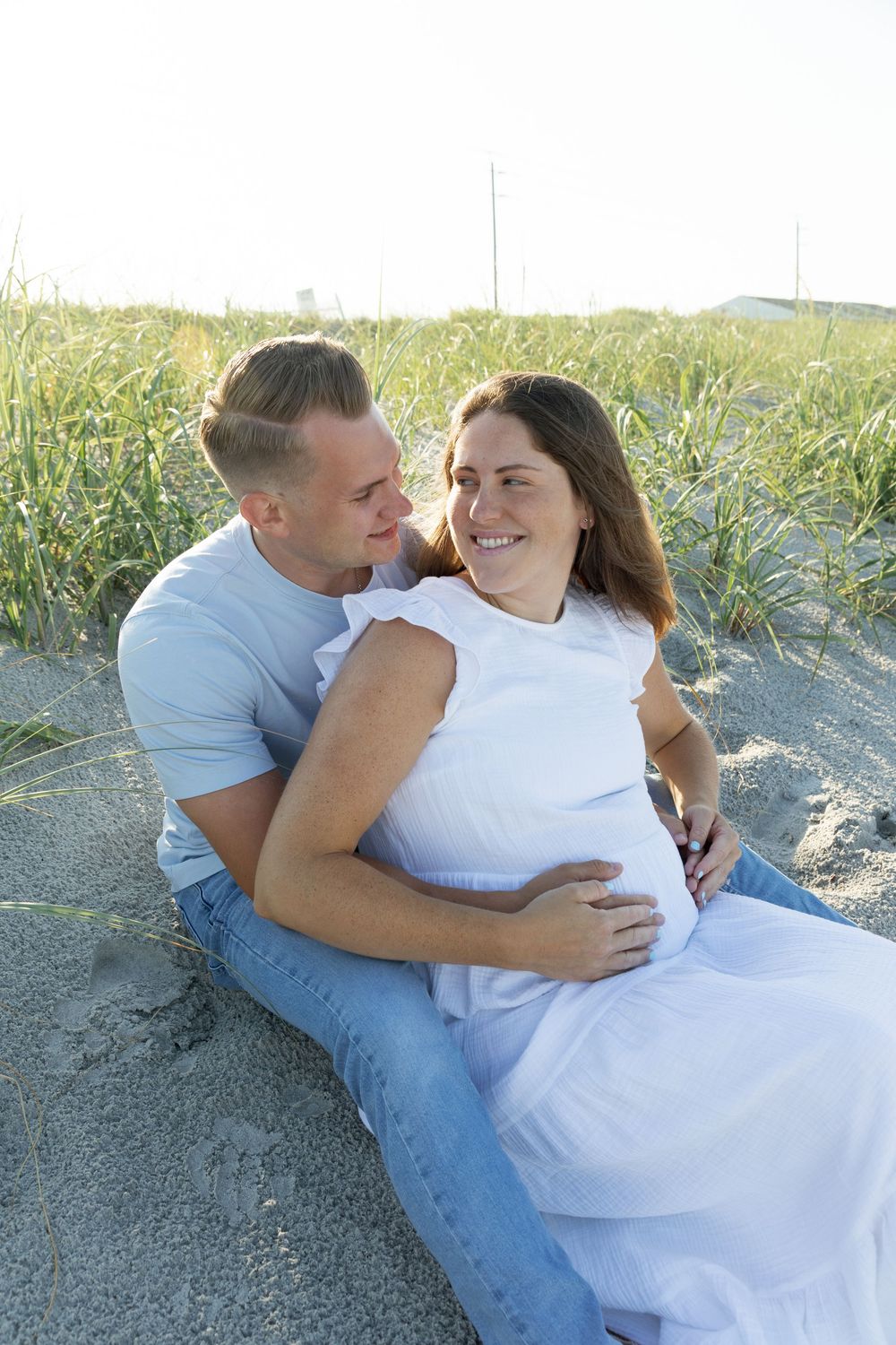 Couple sitting on beach sand dunes, pregnant woman in white dress smiling.