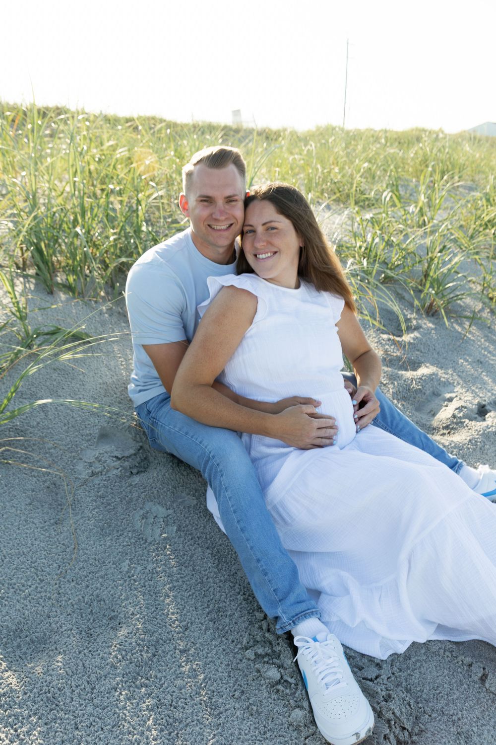 Happy couple sitting on sandy beach, pregnant woman in white dress embracing.