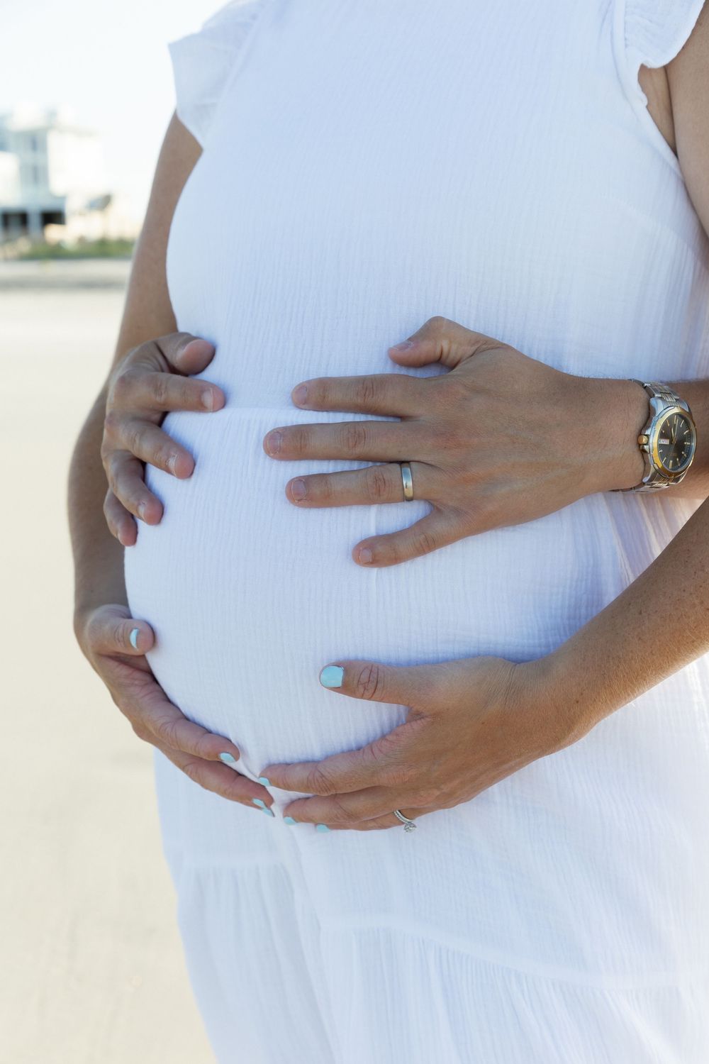 Close-up of pregnant woman in white dress cradling baby bump with both hands.