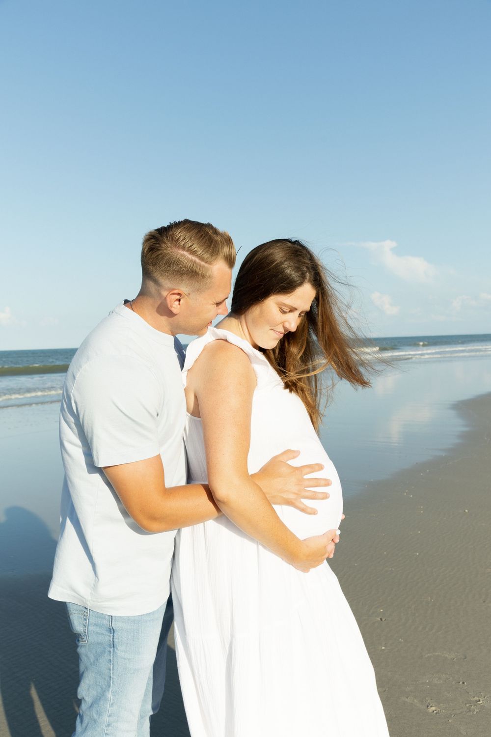 Couple standing on beach, man embracing pregnant woman in flowing white dress.