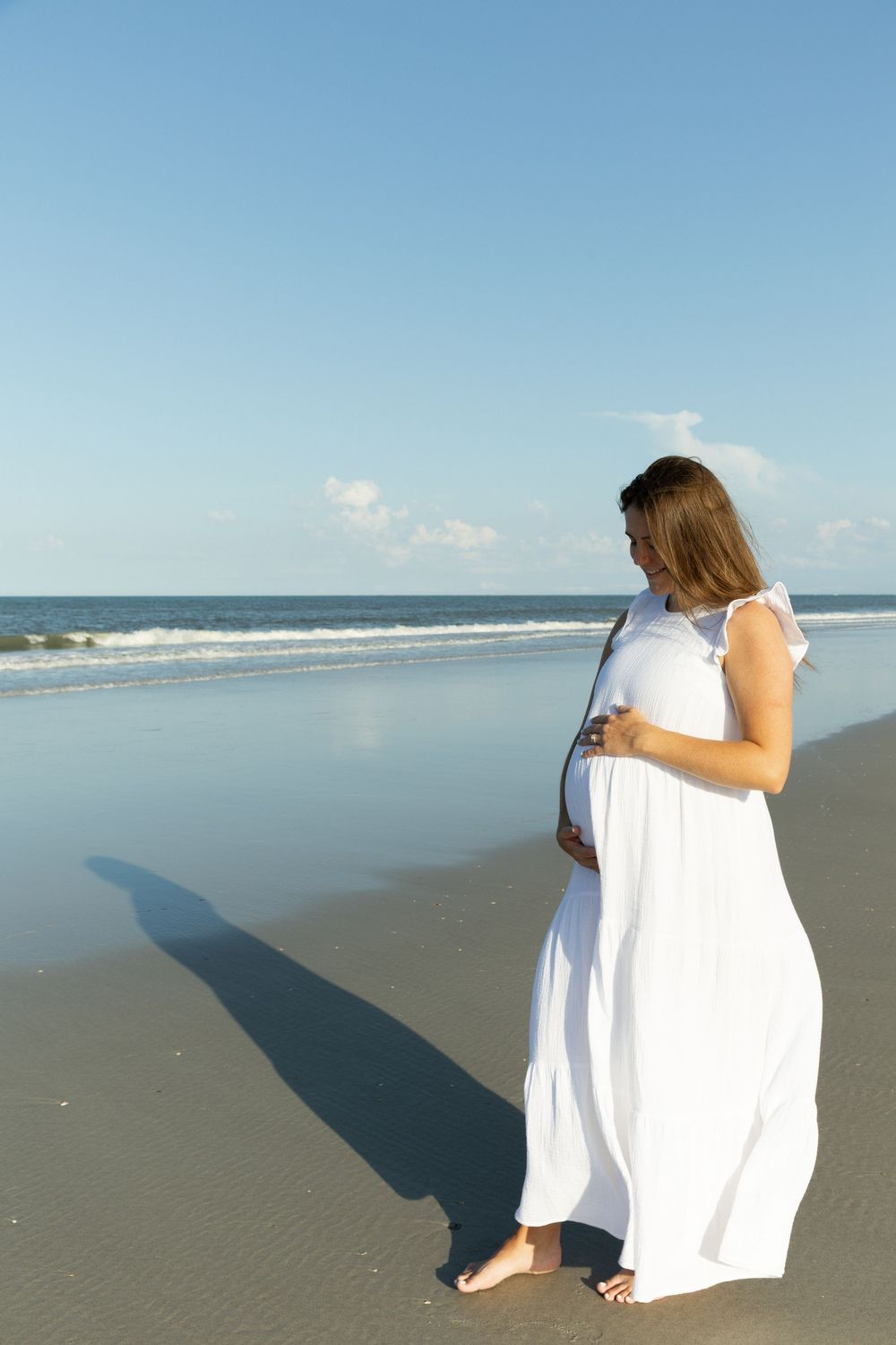 Pregnant woman in white maxi dress standing on beach holding her baby bump.