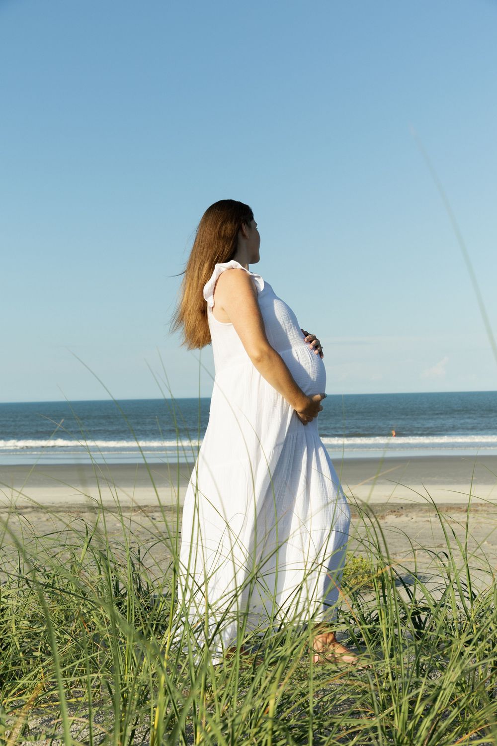 Pregnant woman in white dress standing on beach dunes, holding belly while gazing at ocean.