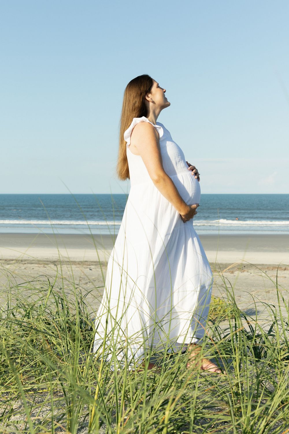 Pregnant woman in white dress standing in beach grass gazing at the ocean.