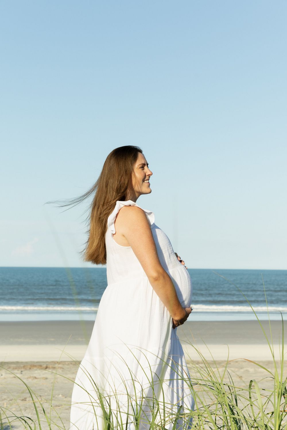Smiling pregnant woman in white dress standing near beach grass with ocean in background.