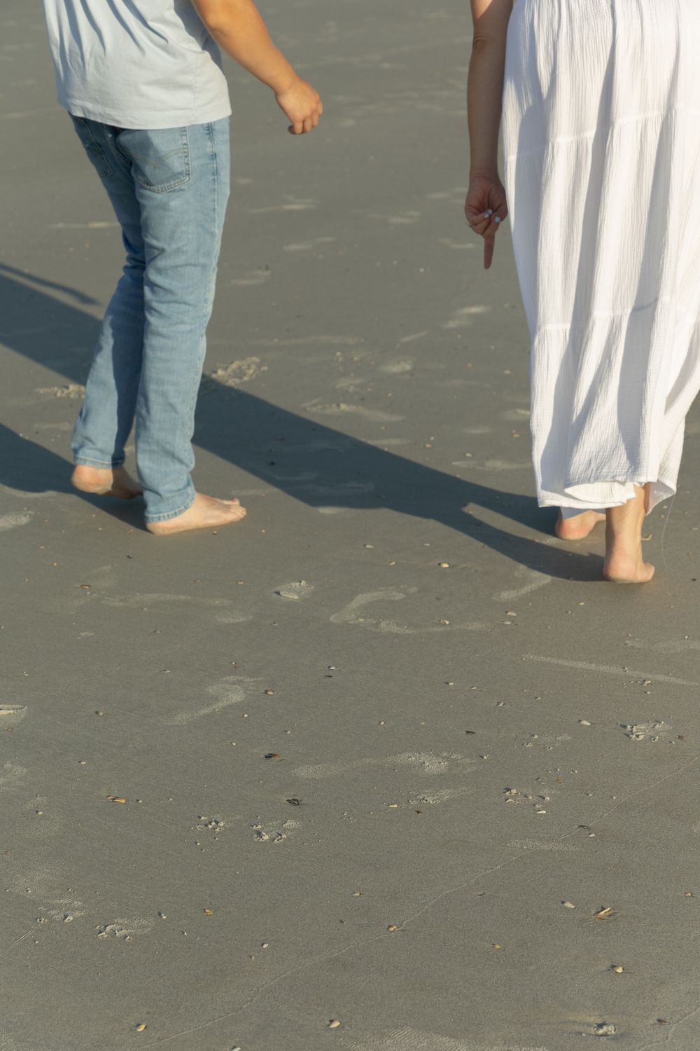 Barefoot couple walking on sandy beach, man in jeans and woman in white dress.