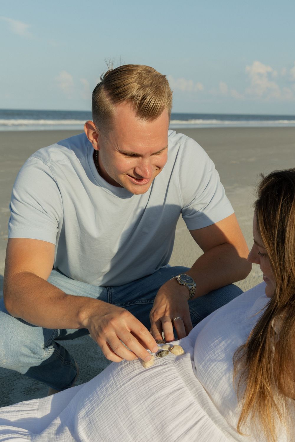 Man sitting on beach showing seashells to pregnant woman during golden hour sunset.