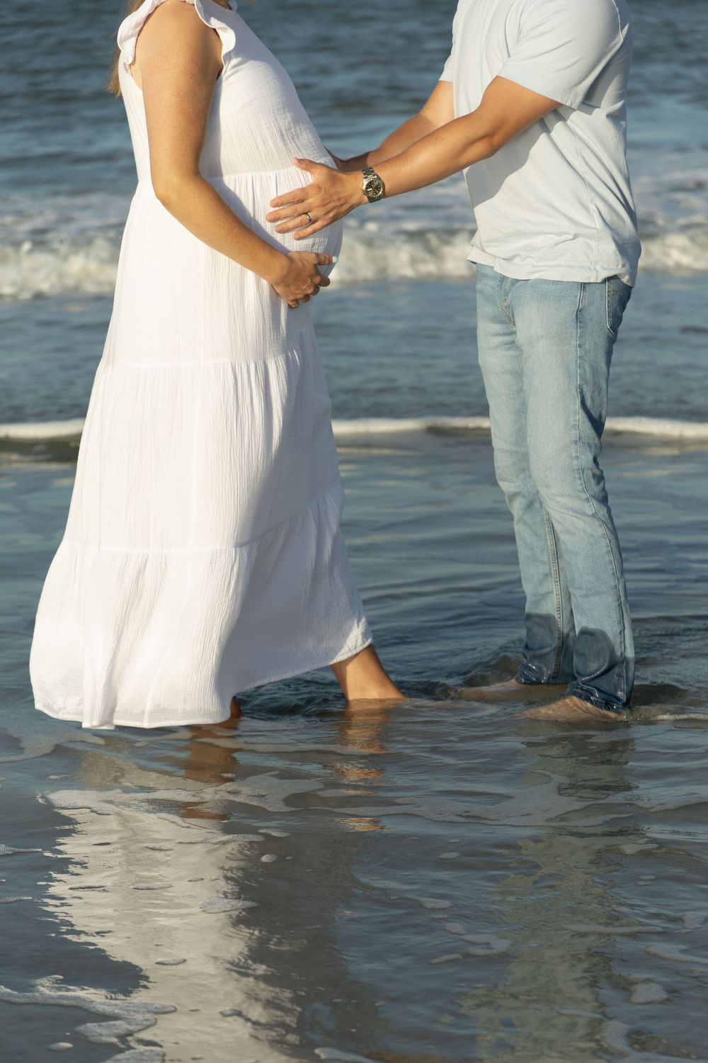 Couple standing in ocean waves, man holding pregnant womans belly on sunny beach.
