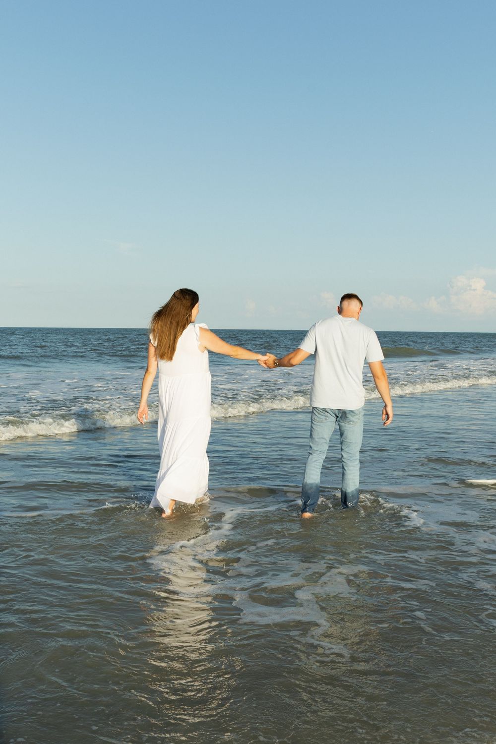 Pregnant couple holding hands walking into ocean waves on a clear sunny day.