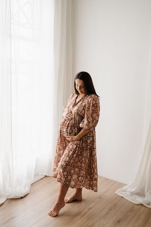 Pregnant woman in floral dress poses by window during maternity photoshoot.