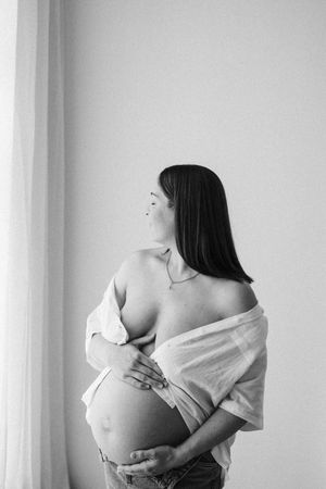 Artistic black and white maternity photo of woman wearing loose shirt by window.