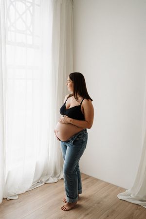 Pregnant woman in black top and jeans gazes out window during intimate photoshoot.