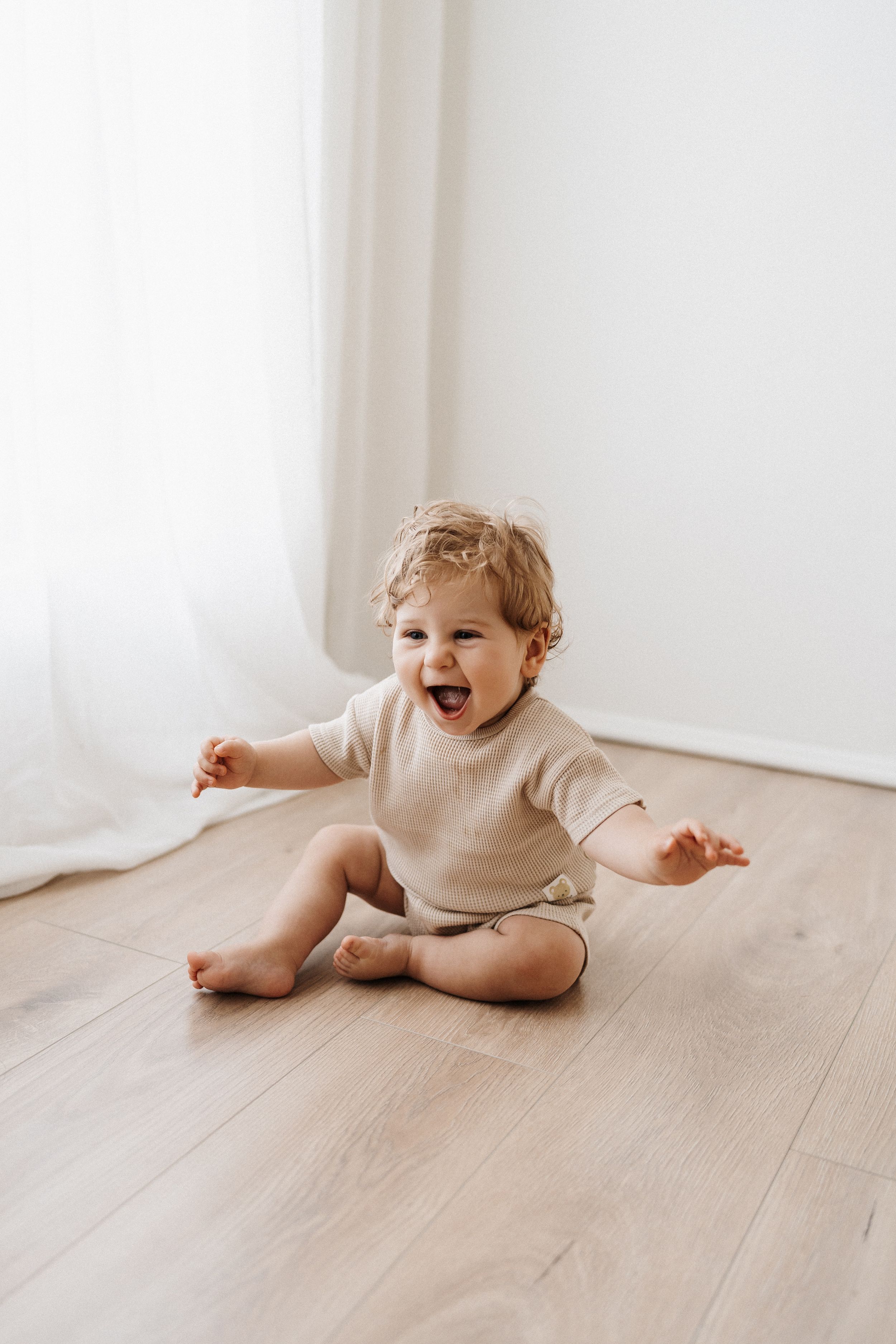 Happy toddler in beige sweater laughing and playing on wooden floor in front of white curtains.