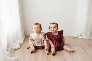 Twin babies in matching ruffled outfits sitting together on white floor.