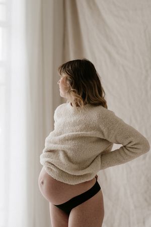 Side view of pregnant woman in cozy beige sweater and black underwear.