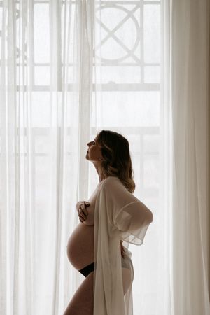 Silhouette of pregnant woman in sheer white robe against bright window.
