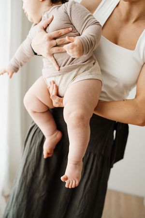 Parent holding baby wearing beige onesie showing chubby legs and bare feet against white wall.