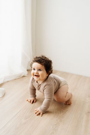 Joyful baby in beige onesie crawling on wooden floor near white curtain.