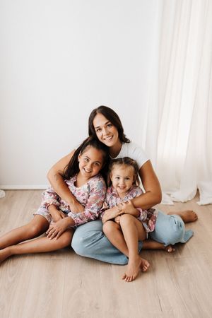 Mother and two young daughters in matching floral dresses sitting together on wooden floor.