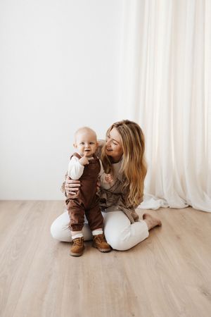 Parent and toddler in brown overalls share a tender moment by white curtains on wooden floor.