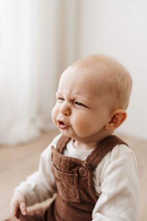 Baby in brown overalls and white shirt making amusing grumpy expression.