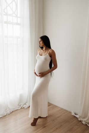 Expectant mother in white dress poses elegantly by window during maternity photoshoot.