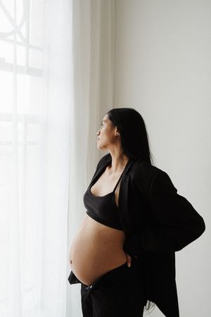 Pregnant woman in black clothing stands by window looking to side in minimalist maternity photo shoot.