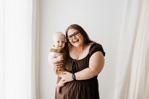 Woman in black dress holding baby in brown outfit against white background.