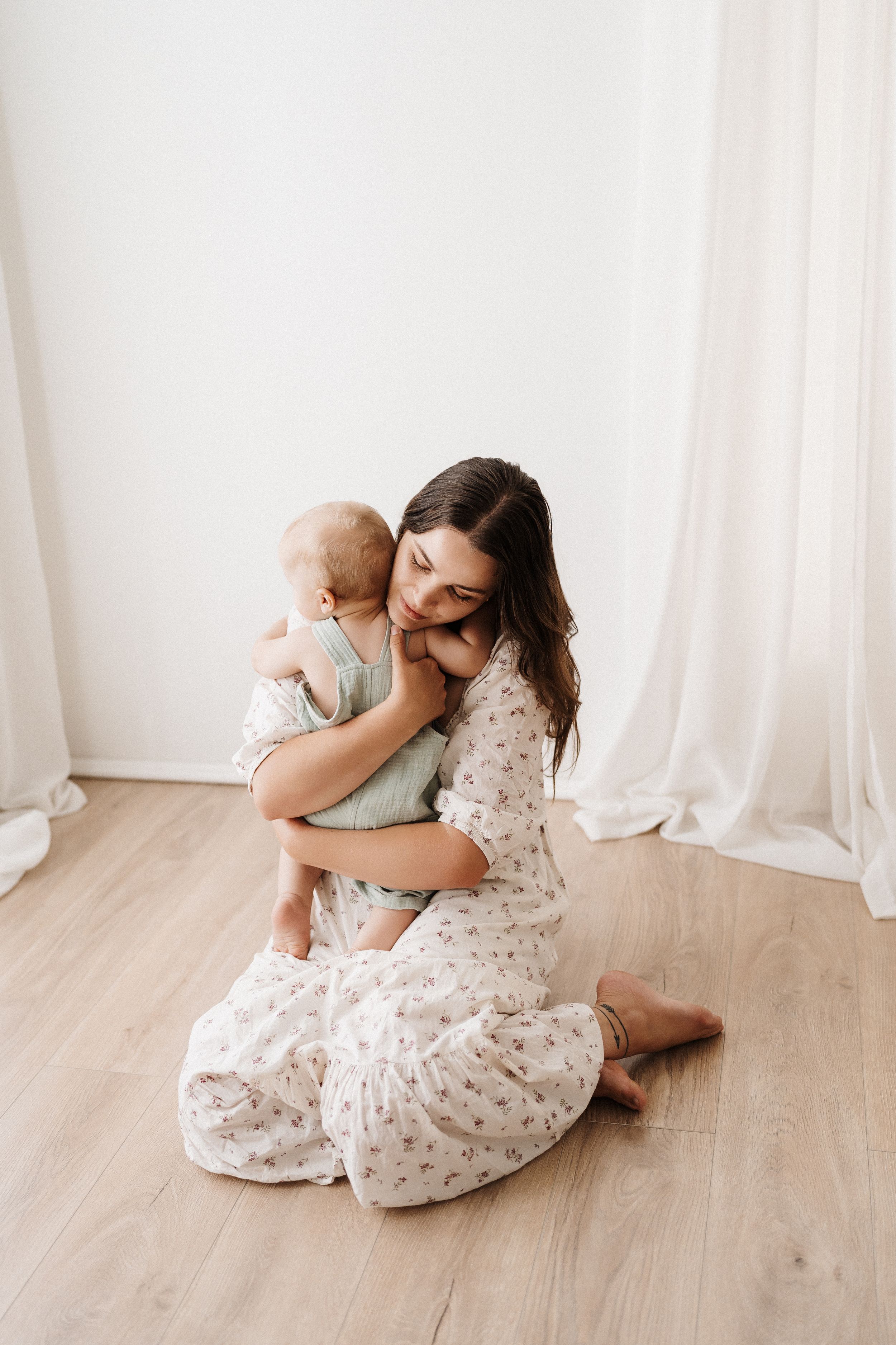 A tender embrace against white curtains and light wood flooring creates an intimate moment in soft natural light.