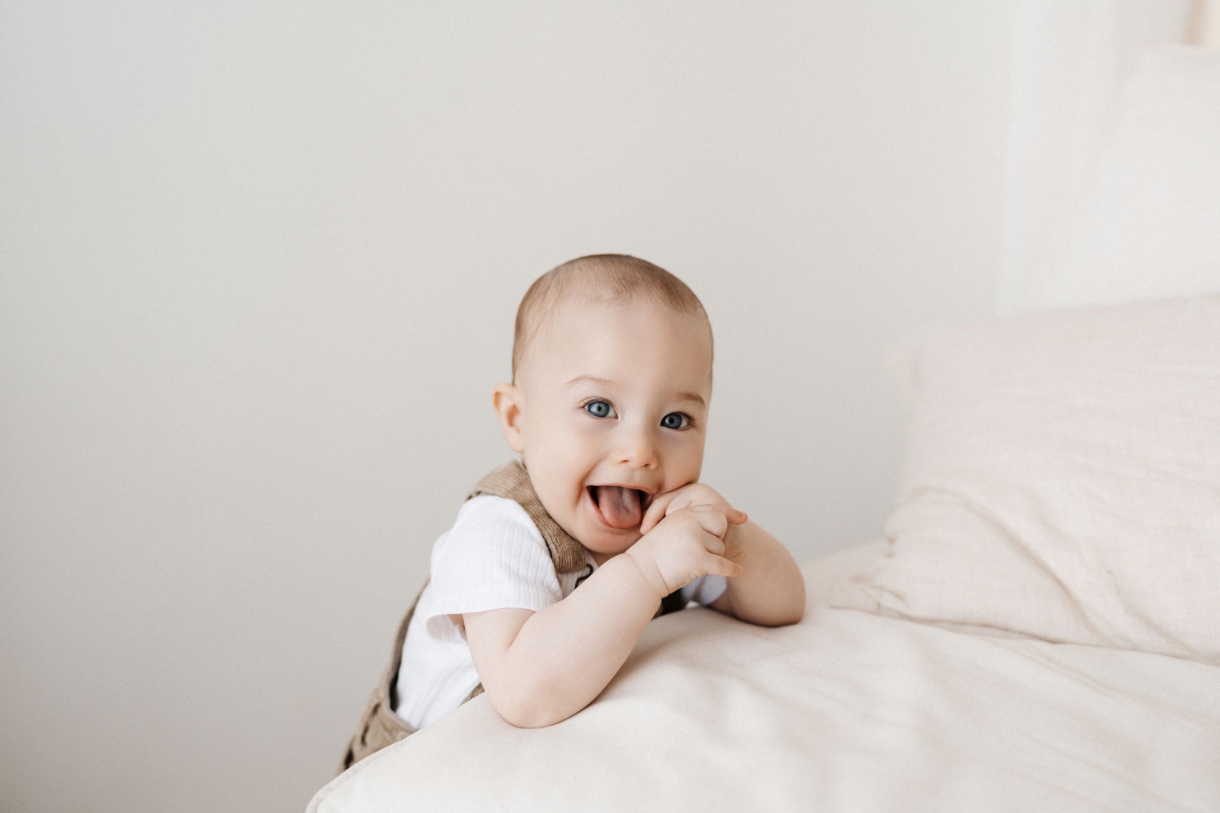 Baby in white onesie playing and laughing on light colored bedding.