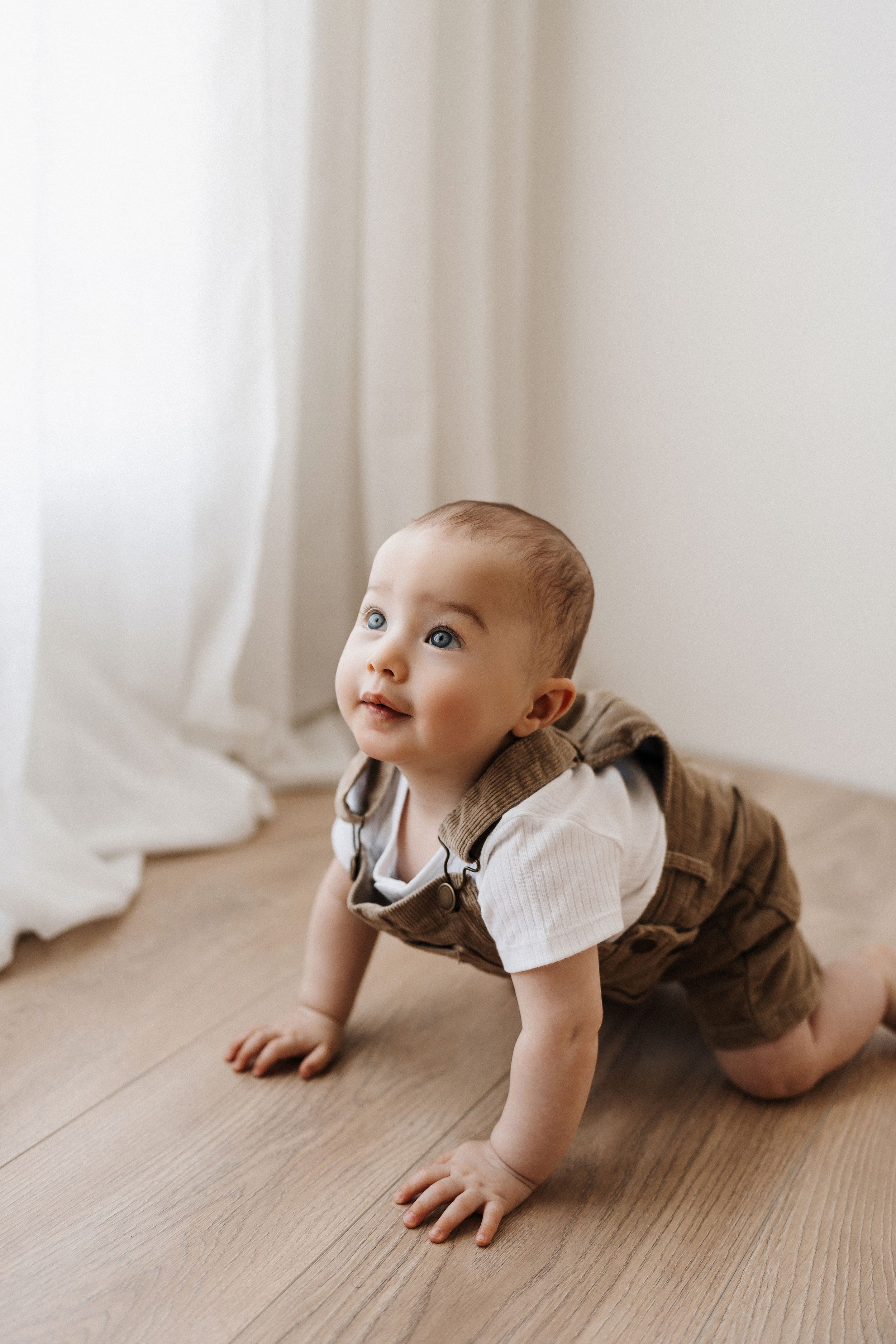 Baby crawling on wooden floor wearing white shirt and brown pants.