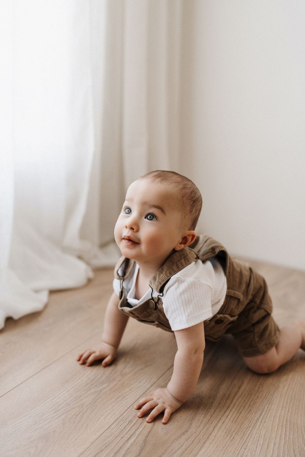 Baby crawling on wooden floor wearing white shirt and brown pants.