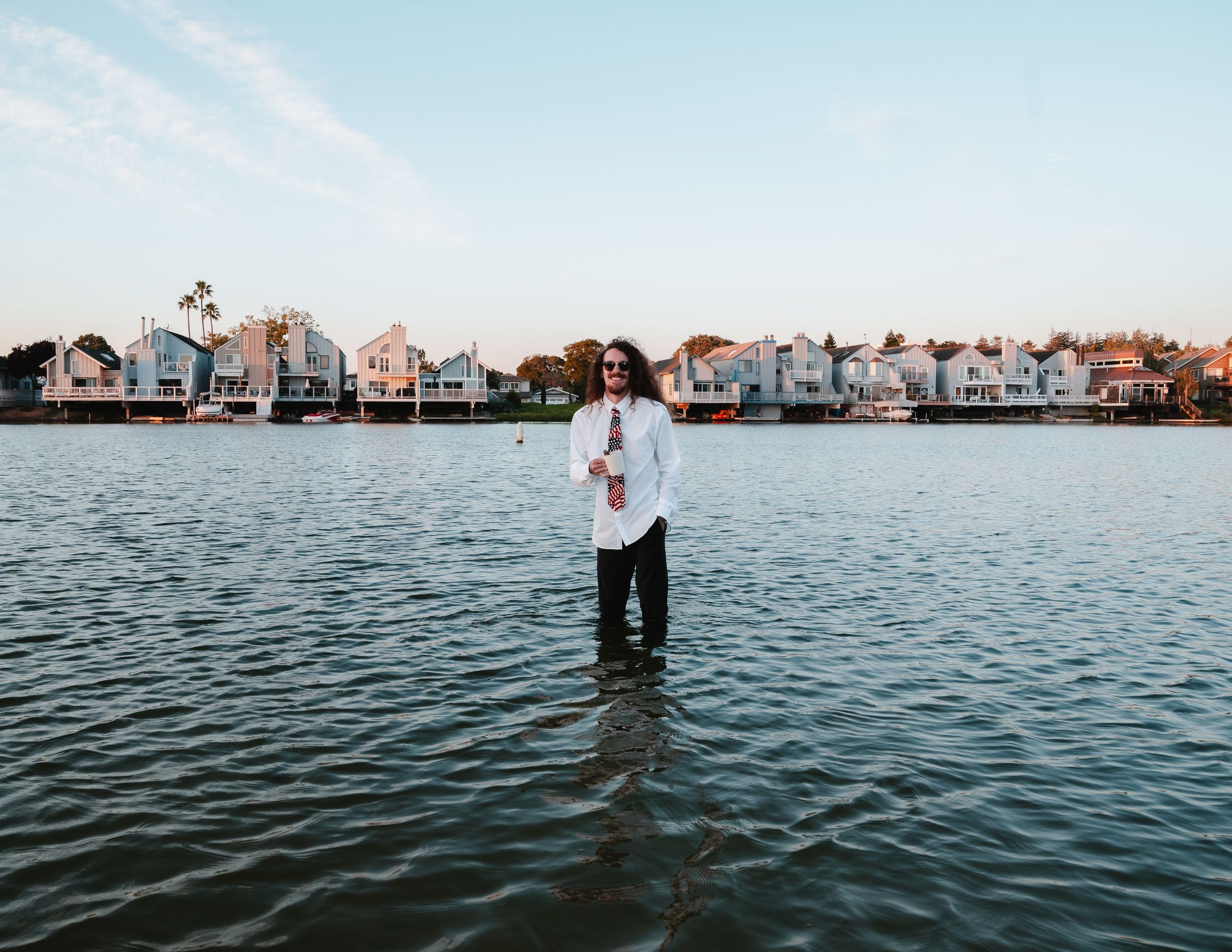Person standing in coastal waters with upscale waterfront houses and boats visible along the shoreline at sunset.