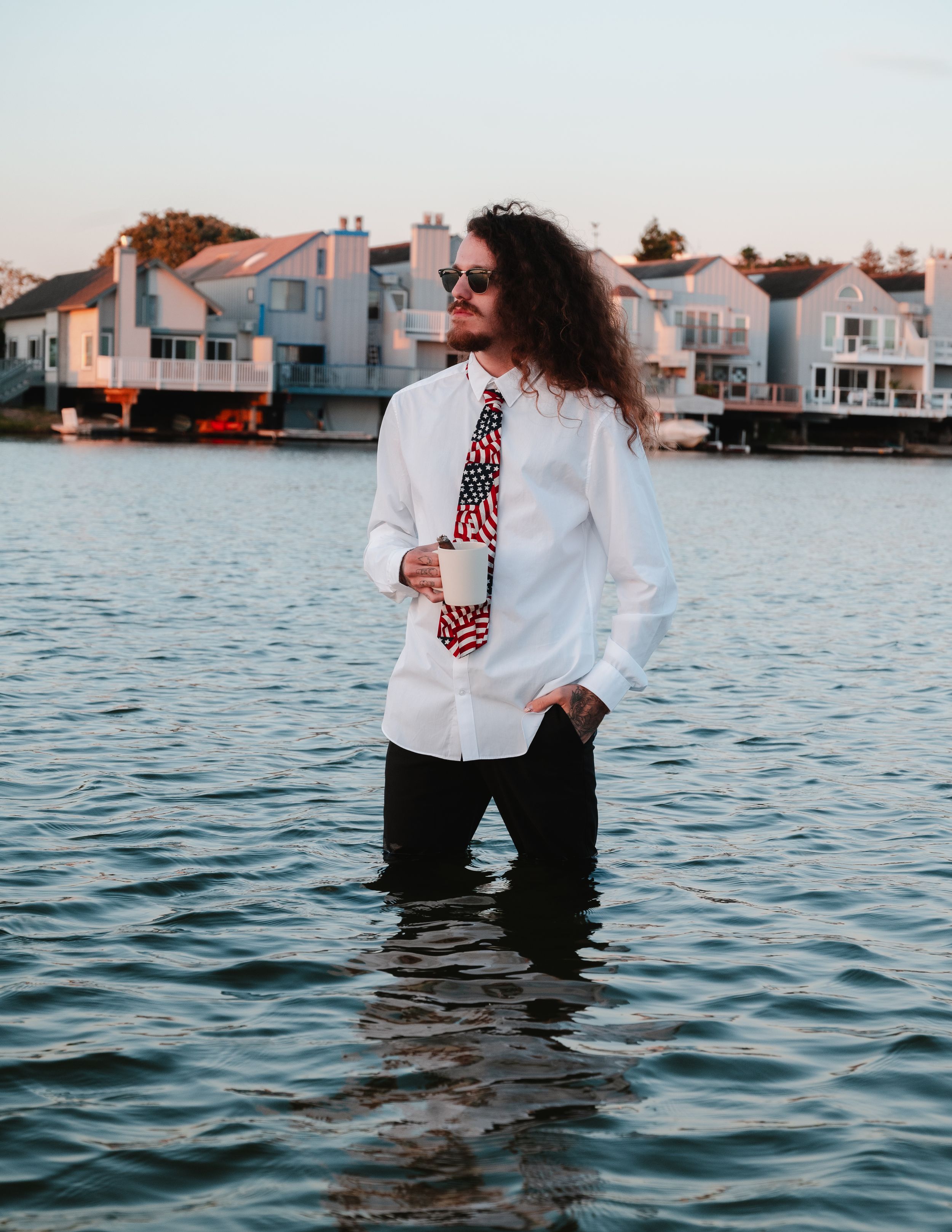A couple wearing black and white attire stand in shallow water at sunset near waterfront homes.