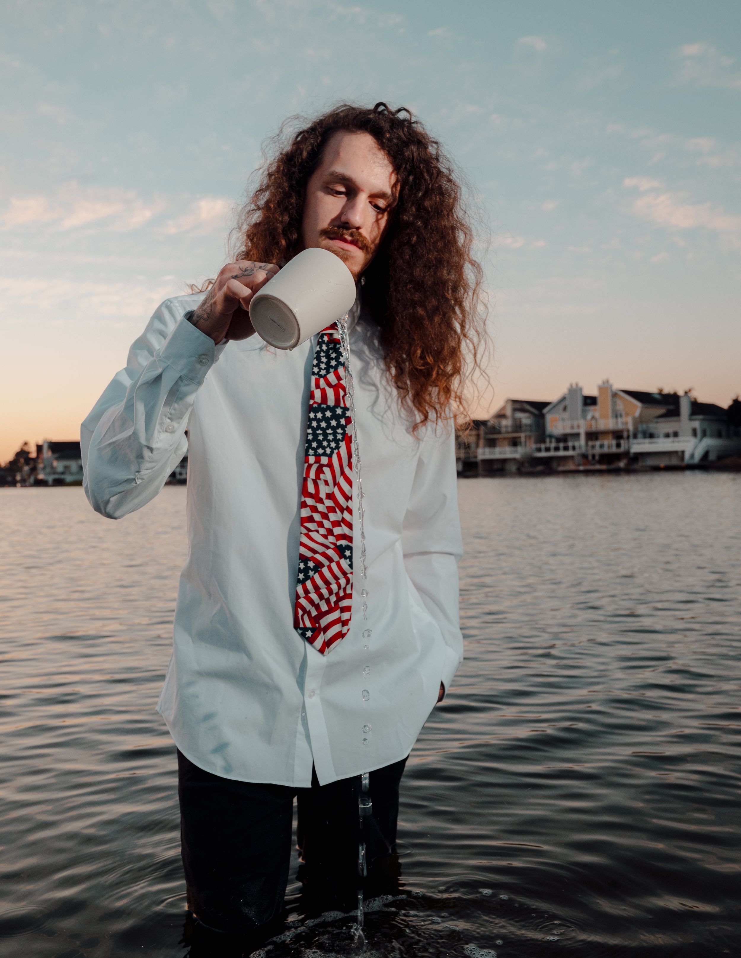 Person in white shirt with patriotic tie drinks from cup against waterfront sunset backdrop.