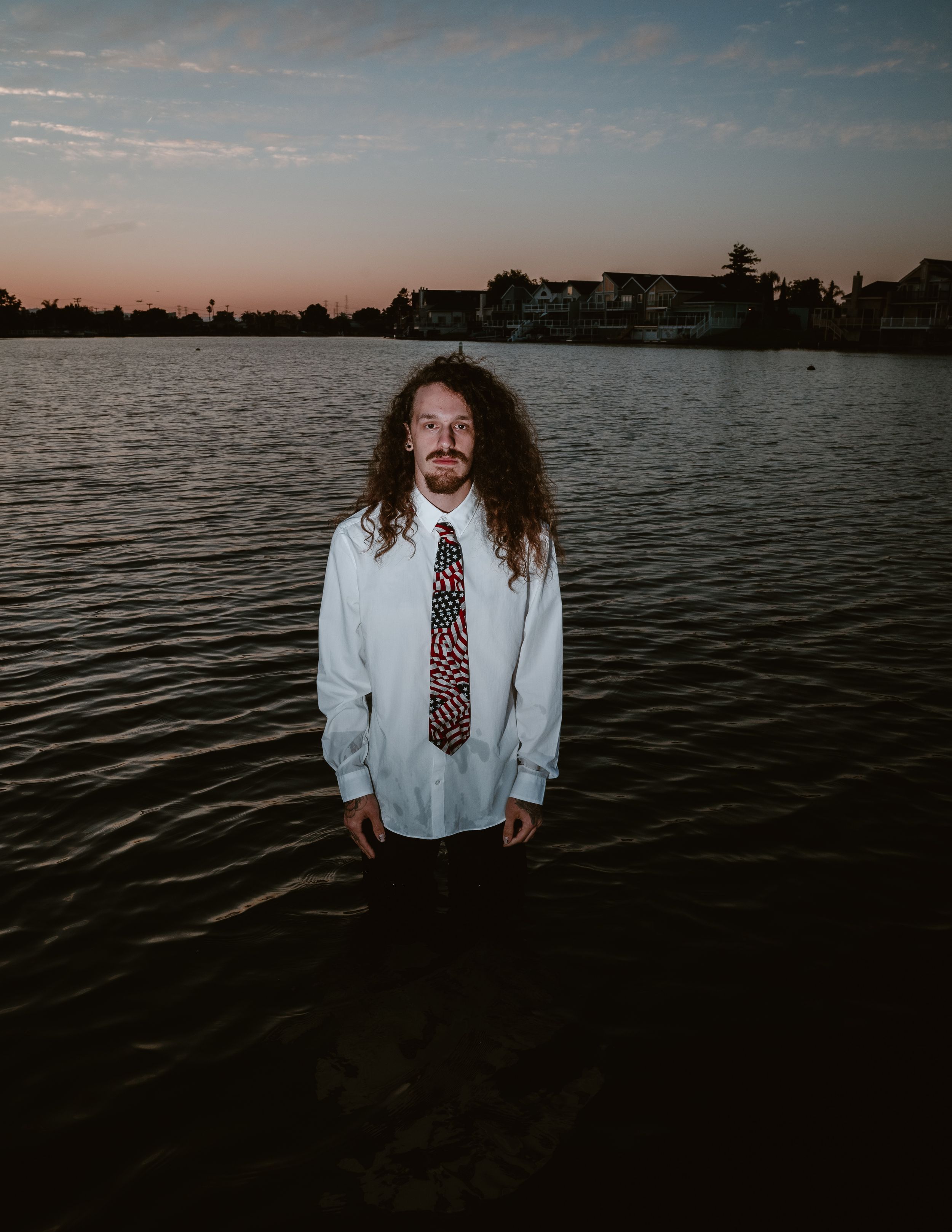 Figure stands in white shirt and patterned tie against lake at sunset with houses visible on distant shore.